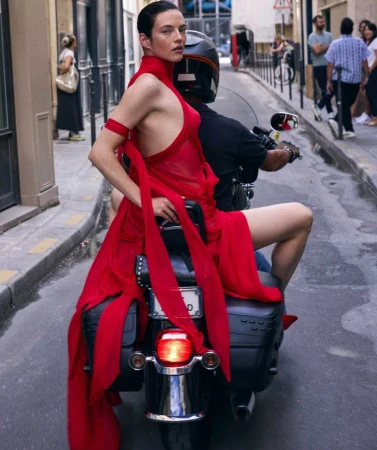 Fashionable woman in red gown riding a motorcycle through a European city street, showcasing style and urban mobility.