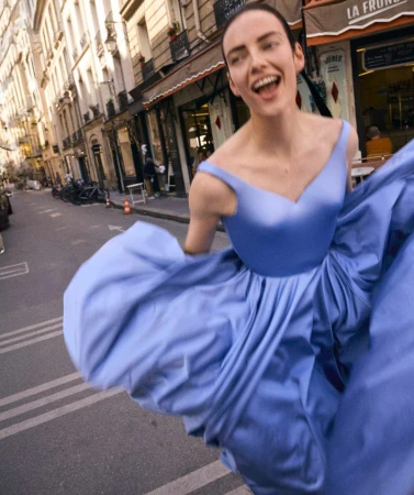 A happy woman in a blue gown runs down a Paris street lined with buildings and shops on a sunny day. Fashion, lifestyle concept.