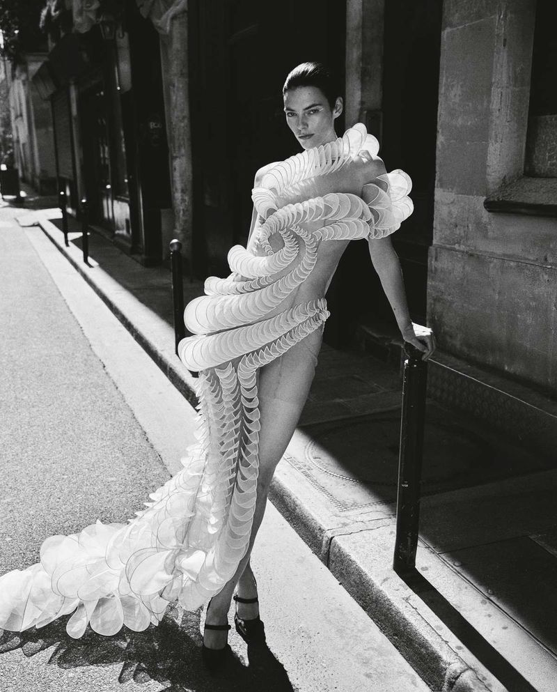 Black and white portrait of a woman in an elaborate white ruffled dress posing on a city street next to a metal railing.