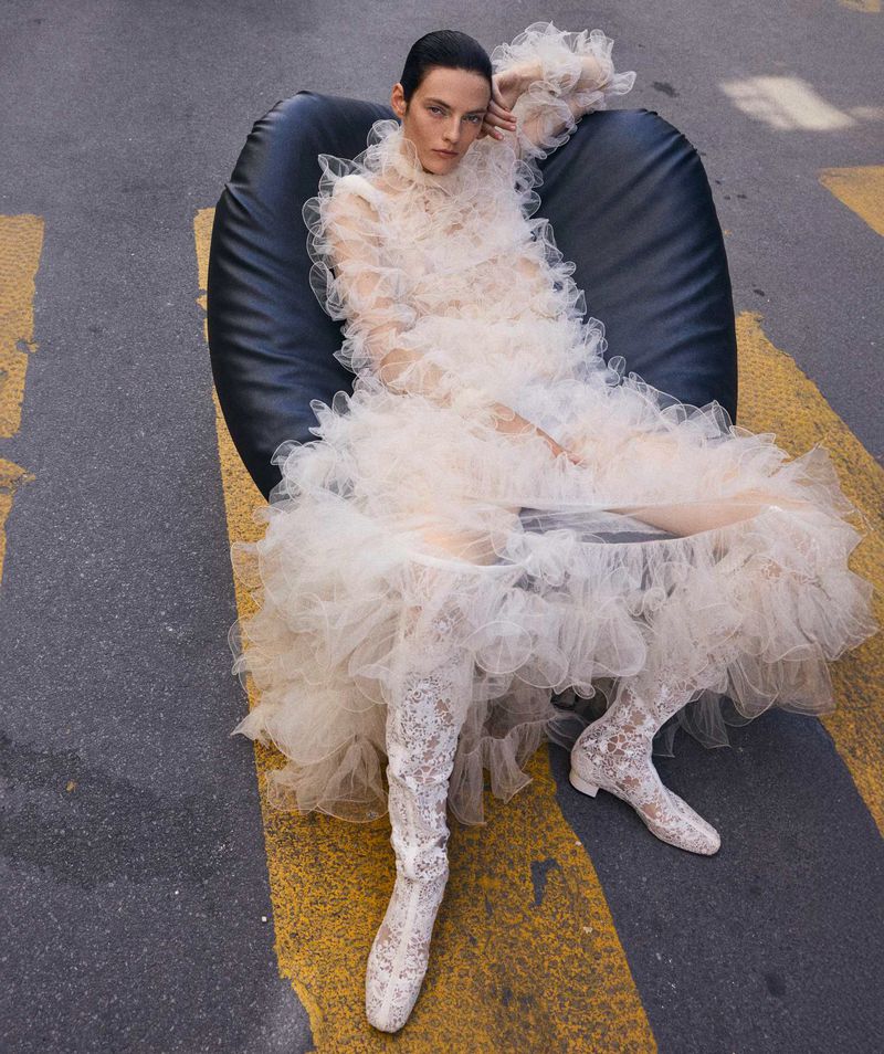 Adult female in a voluminous ruffled dress, reclined in a black chair, on a street crossing with yellow stripes and grey asphalt.