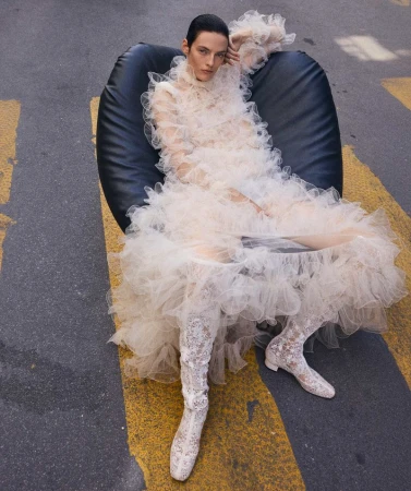 Adult female in a voluminous ruffled dress, reclined in a black chair, on a street crossing with yellow stripes and grey asphalt.