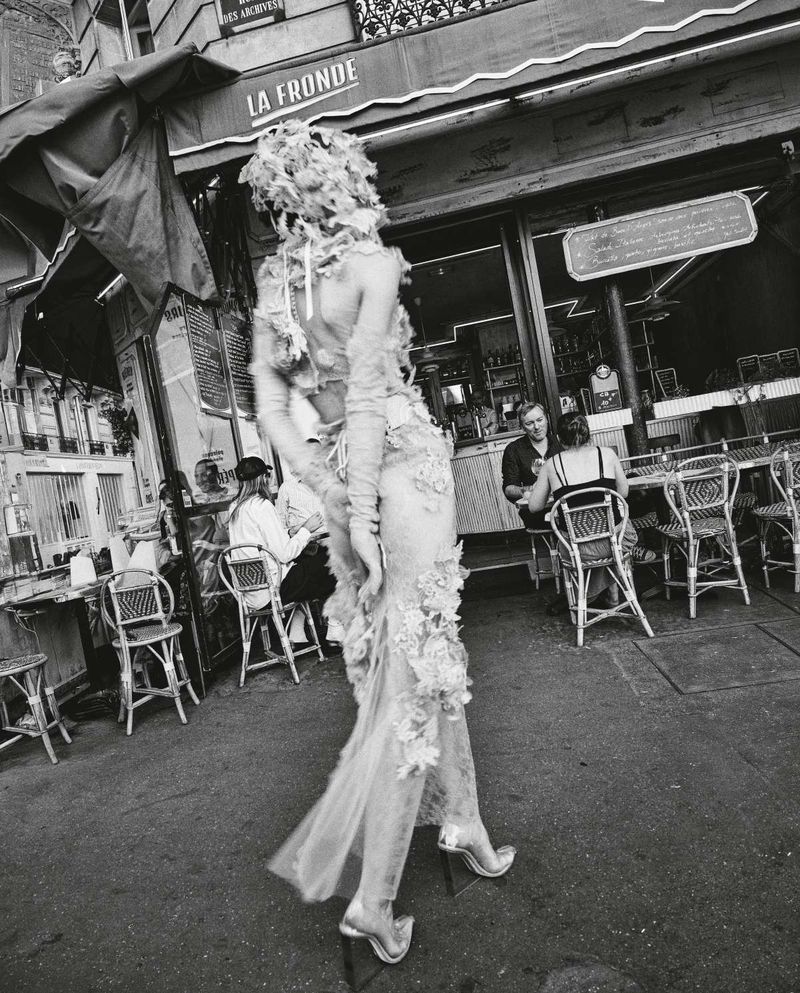 Monochrome shot of a person in an avant-garde dress walking past a cafe, capturing a blend of high fashion and everyday urban life.