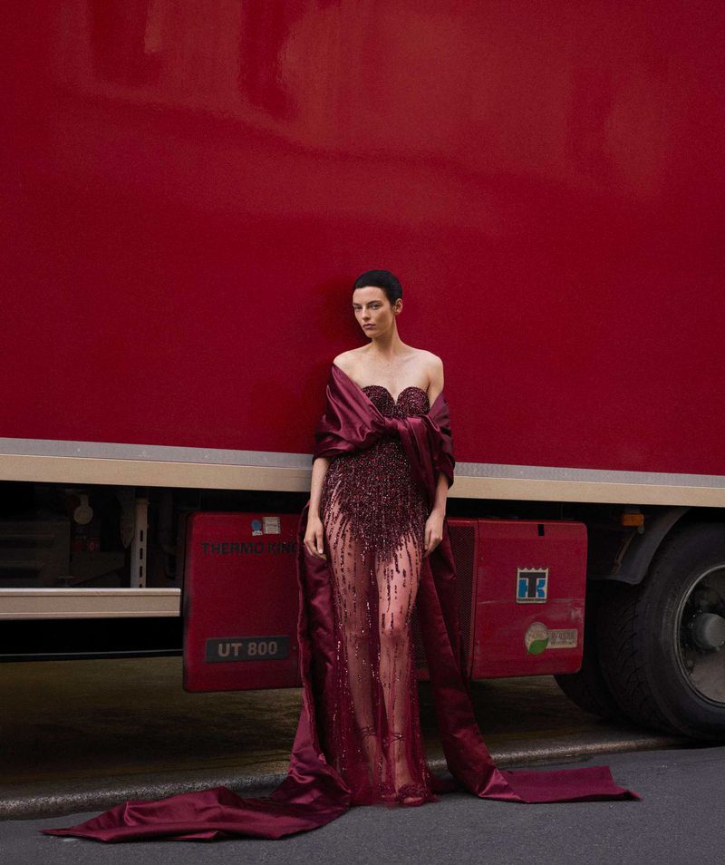 Woman poses in a dark red gown with a train in front of a solid-red truck. Fashion photography with natural light and street setting.