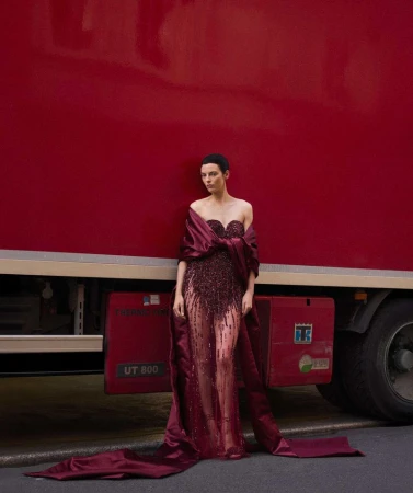 Woman poses in a dark red gown with a train in front of a solid-red truck. Fashion photography with natural light and street setting.