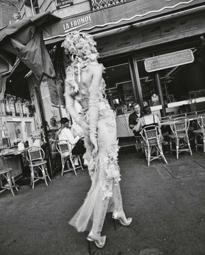 Model in textured sheer gown walking past an outdoor cafe with patrons