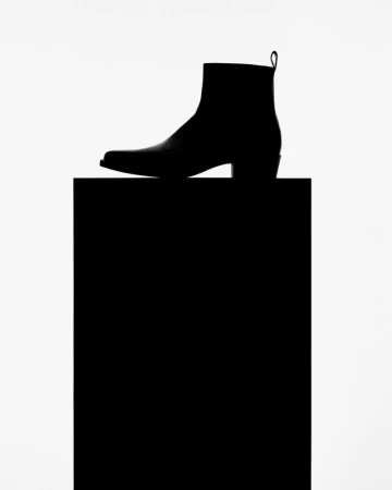 Black leather heeled boot stands on a black block against a white background in a studio shot for a minimalist fashion advertisement.