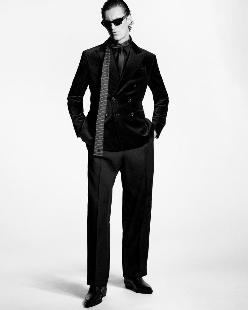 A full-length black and white shot of a male model wearing a black velvet suit, dark shirt, tie, sunglasses, and black leather boots.