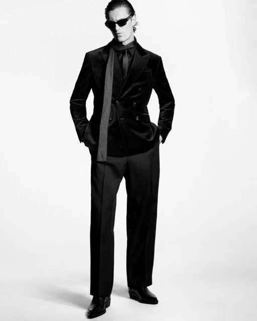 A full-length black and white shot of a male model wearing a black velvet suit, dark shirt, tie, sunglasses, and black leather boots.