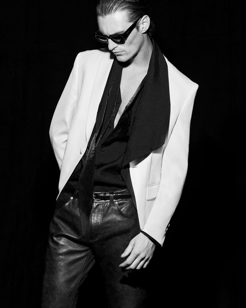 Monochrome portrait of a stylish adult male in a blazer and sunglasses, looking down against a dark backdrop in a fashion-editorial style.