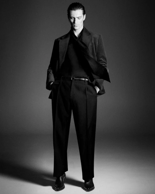 Full-length studio shot of an adult male in a suit, hands in pockets, wearing a black top and dark trousers, standing on a grey backdrop.