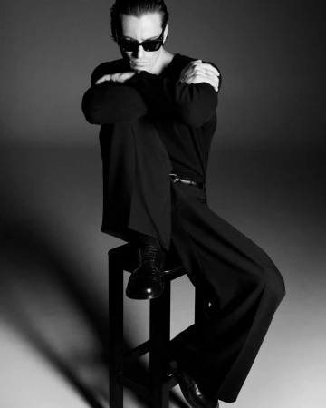 Man wearing dark sunglasses and black long-sleeve shirt seated on a stool against a grey studio backdrop