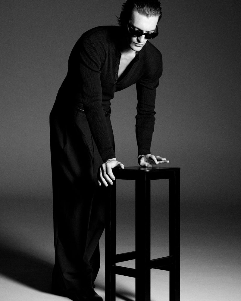 Monochrome fashion portrait of a stylish adult male leaning on a black stool in a photo studio with dark sunglasses.
