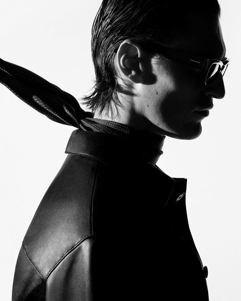 Model Myles Crosby in profile wearing a black leather jacket, dark glasses, and a trailing neck scarf against a stark white backdrop.