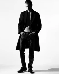 Monochrome full-length portrait of Myles Crosby wearing an open black overcoat and leather trousers against a white background.
