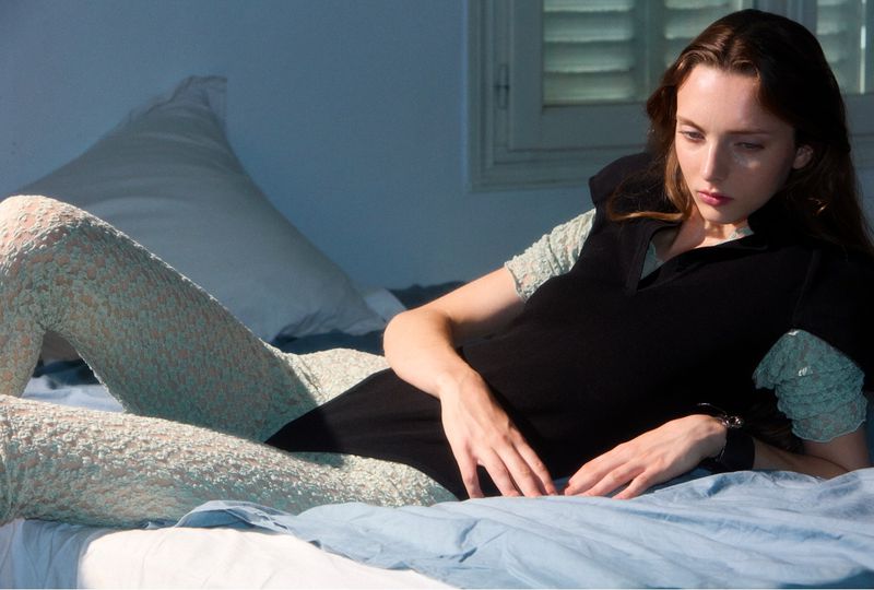A fair-skinned young woman with long brown hair lies on a light blue bed with a white pillow and a black top and patterned leggings.