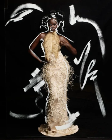 Elegant dark-skinned woman in feathered gown poses against a black backdrop with abstract white paint strokes for a high fashion editorial.