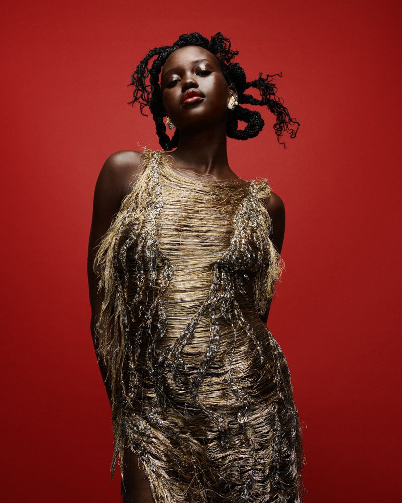 Dark-skinned woman in textured gold dress poses against red background. Shoulder-length braided hair frames face in an indoor studio setting.