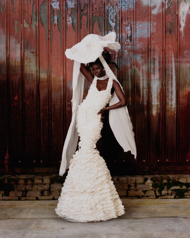 Elegant African woman in a white ruffled wedding dress and pearl jewelry poses in front of a rustic, distressed metal and brick wall.
