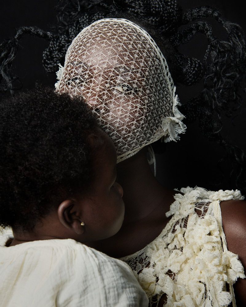A mother holds her baby, both with dark skin tones, in a portrait on a black background. The mother wears a textured veil and dress.