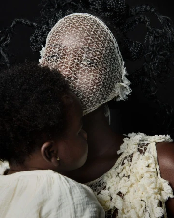 A mother holds her baby, both with dark skin tones, in a portrait on a black background. The mother wears a textured veil and dress.