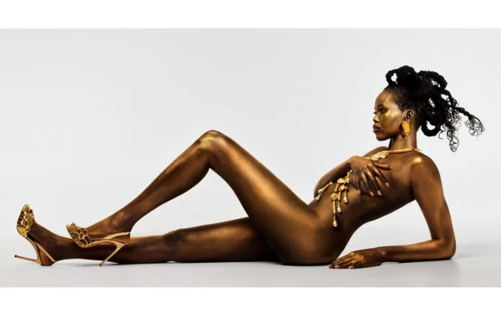 Nude woman coated in metallic gold with golden jewelry and heeled sandals posing on a white background in a glamour-style image.