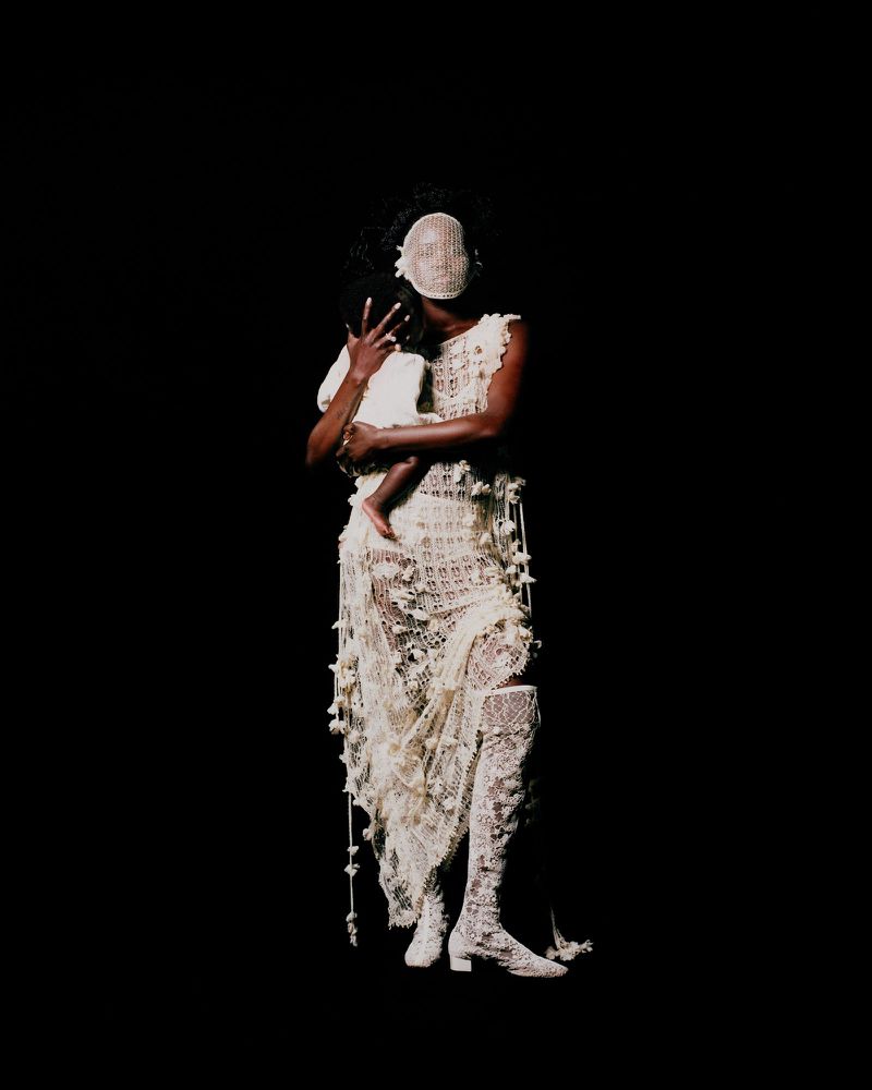 Fashion shot of a woman in an elaborate, textured dress holding a child against a dark background, highlighting unique garment design.