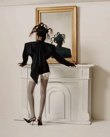 A woman stands in front of a white fireplace and a gold-framed mirror wearing a dark blazer, light stockings, and black heels.