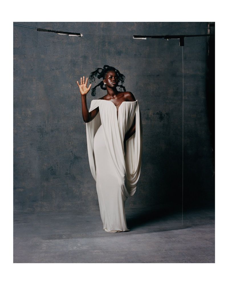 Elegant Black woman in flowing white gown poses in a studio setting with unique hair, striking a stylish pose against a grey backdrop.