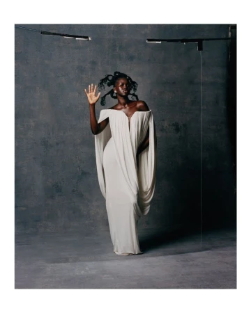 Elegant Black woman in flowing white gown poses in a studio setting with unique hair, striking a stylish pose against a grey backdrop.