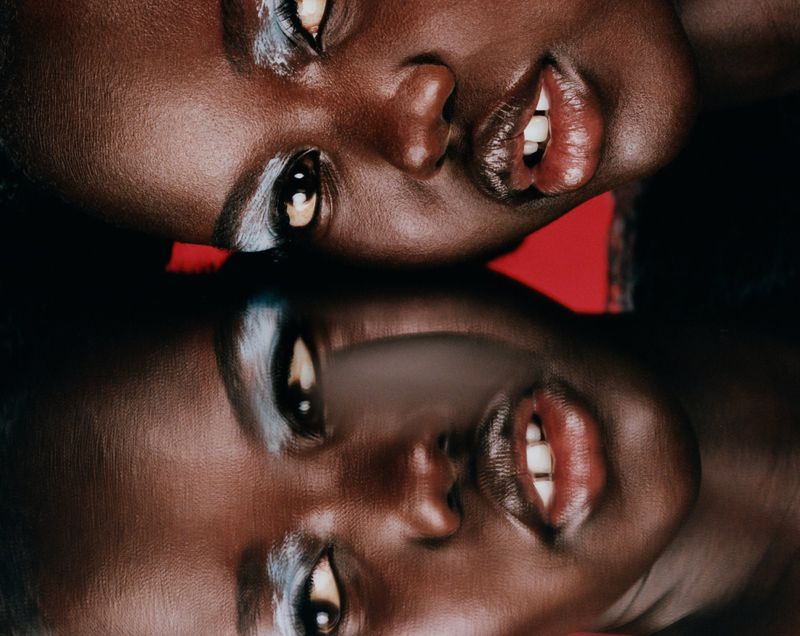 Close-up portrait of an African American woman with silver eyeshadow reflected upside down on a dark reflective surface. The background is red.