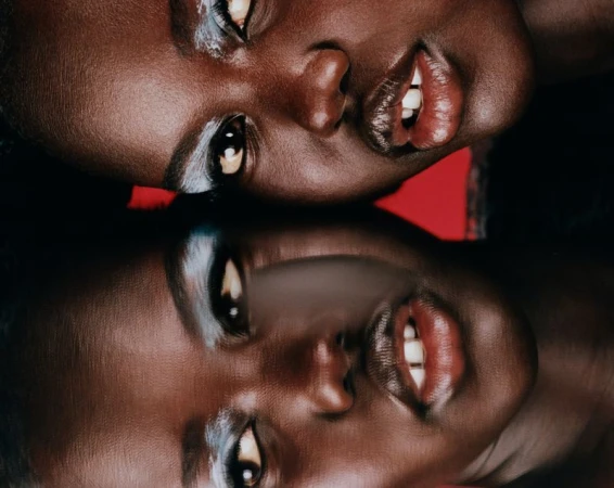Close-up portrait of an African American woman with silver eyeshadow reflected upside down on a dark reflective surface. The background is red.