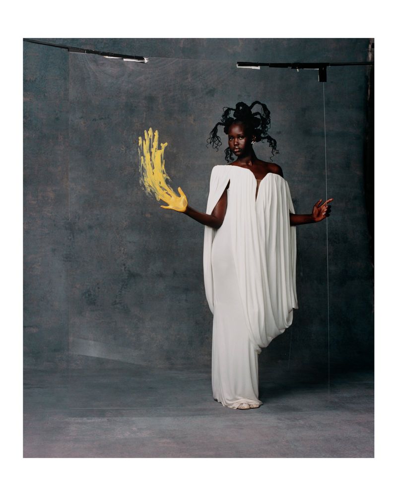 A dark-skinned woman poses in a white dress with yellow paint on her hand in a photo studio against a grey textured backdrop.