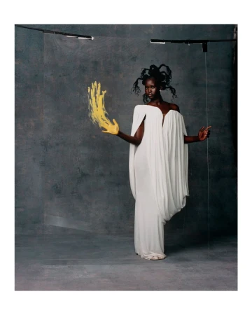 A dark-skinned woman poses in a white dress with yellow paint on her hand in a photo studio against a grey textured backdrop.