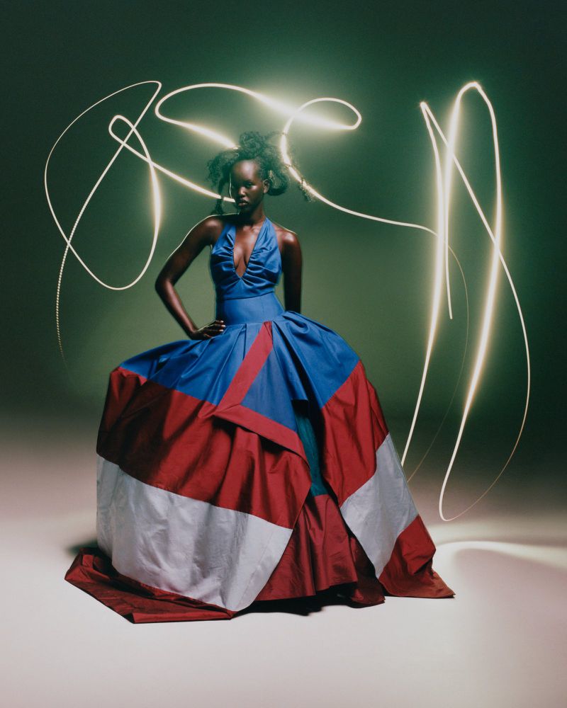 A woman in a colorful dress poses in a studio with light trails. She wears an elegant blue, red, and white gown in a fashion shot.