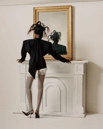 Model seen from behind wearing black blazer, sheer patterned tights, and heels while leaning on a white mantelpiece with a mirror.