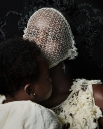 Black woman wearing an intricate cream lace face veil holding a young child against a black studio background.