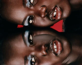 Close-up beauty portrait of a Black woman with glossy lips and silver eyeshadow reflected vertically against a red backdrop.
