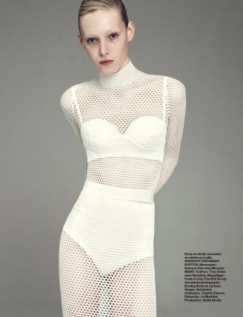 A fair-skinned model in a white mesh dress poses against a light gray backdrop, showcasing a modern fashion editorial style.
