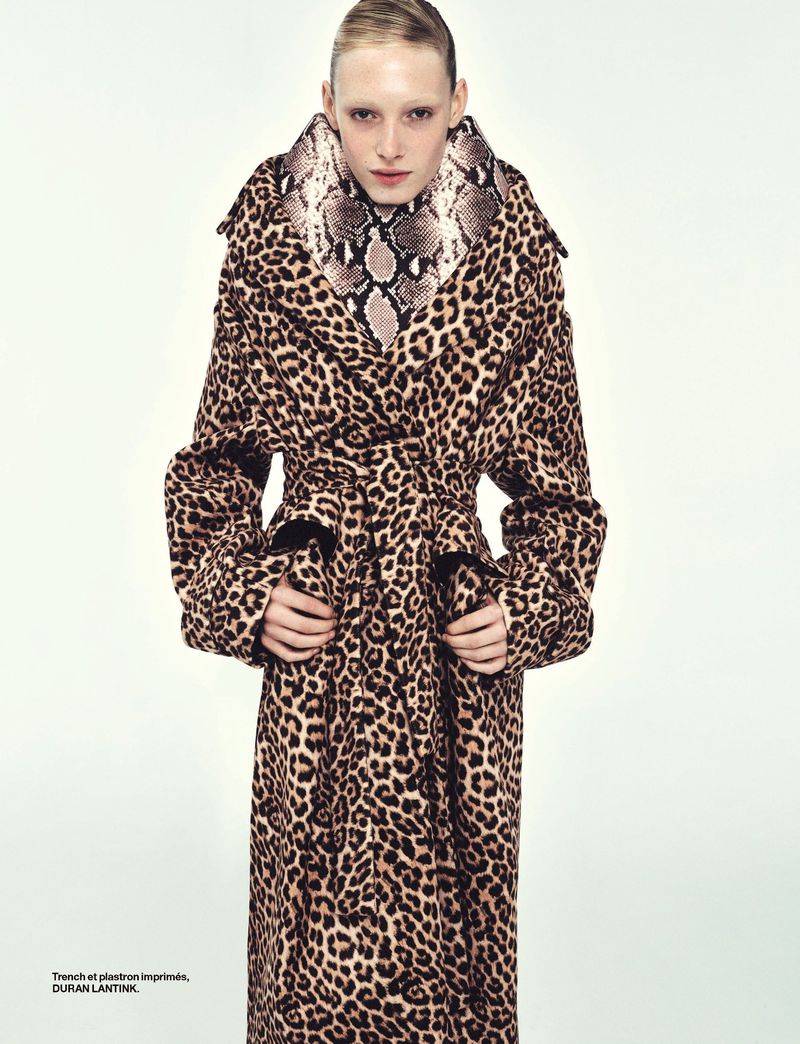 A person with fair skin and light blonde hair wears a long leopard print coat with a snake print collar in a fashion-style studio shot.