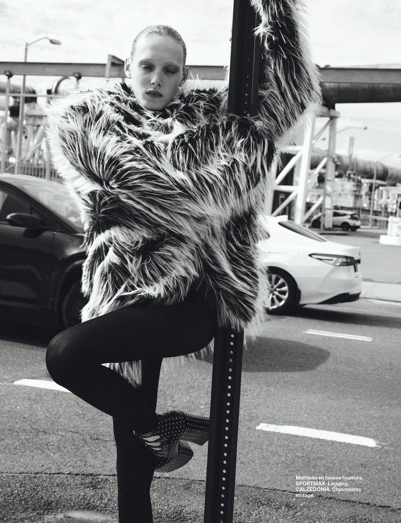 Editorial fashion shot of a woman in a faux fur coat, leaning on a street sign post with cars and industrial backdrop, cool tones.