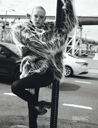 Editorial fashion shot of a woman in a faux fur coat, leaning on a street sign post with cars and industrial backdrop, cool tones.