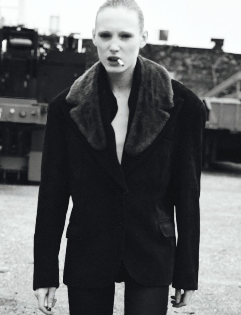 Black and white portrait of a woman in a blazer with fur collar, smoking a cigarette outdoors. Industrial background elements.
