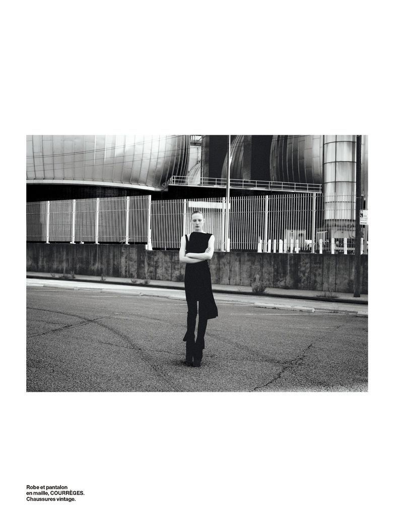 A fashion shot in black and white shows a slender adult female posing in a sleeveless dress in front of an industrial building.