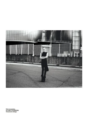 A fashion shot in black and white shows a slender adult female posing in a sleeveless dress in front of an industrial building.