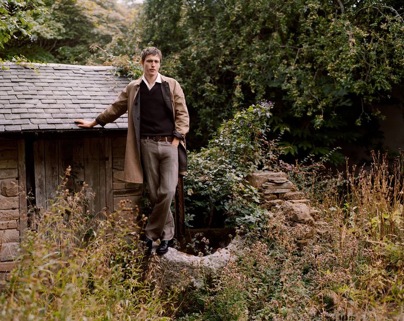 Adult man stands on a stone wall near a stone building. He wears a tan coat, dark shirt, and jeans outdoors in a lush setting.