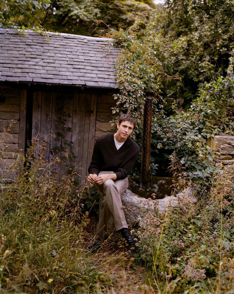 A young adult man sits on a stone slab in front of a rustic building surrounded by lush greenery in a rural outdoor setting.