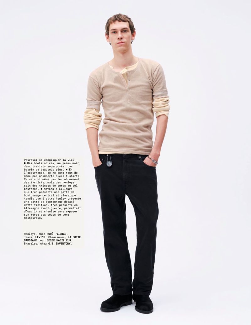 Full-length portrait of a young adult man in a casual beige long-sleeve shirt, black jeans, and dark shoes against a white backdrop.