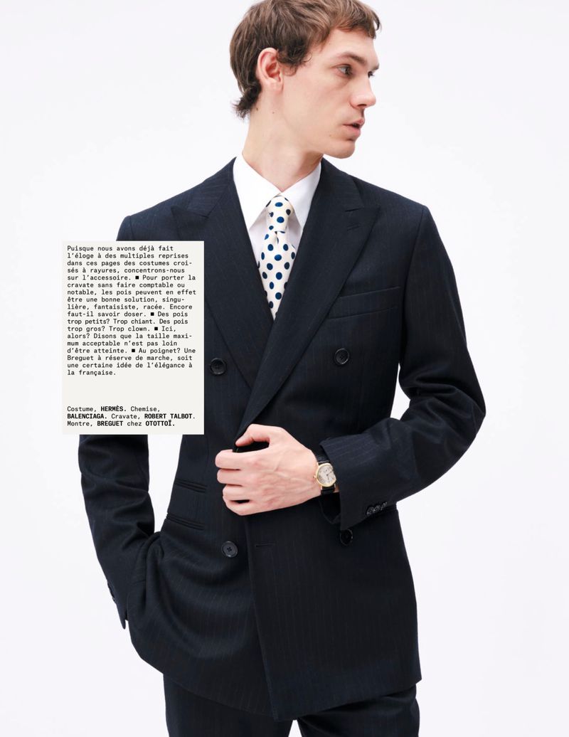 A man in a dark suit and polka dot tie poses against a white background, a printed sheet with text held in front of him.