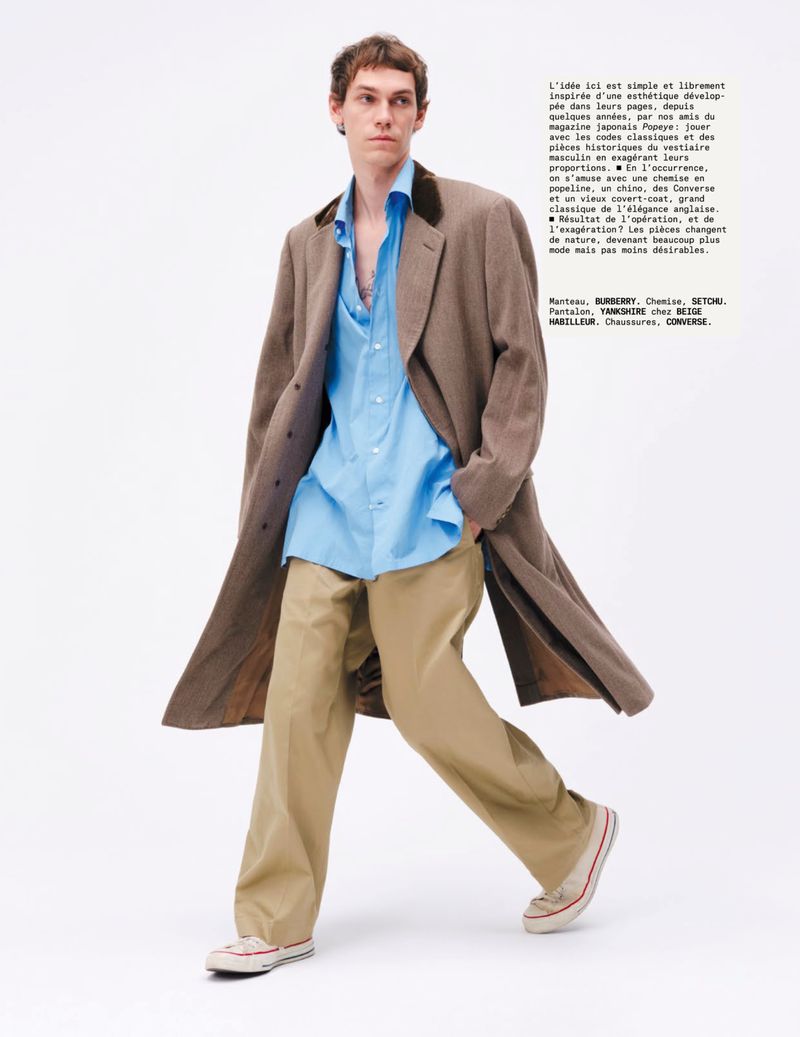 Man in open blue shirt, tan pants, and brown coat walking in studio, wearing Converse sneakers, fashion editorial style against white backdrop.
