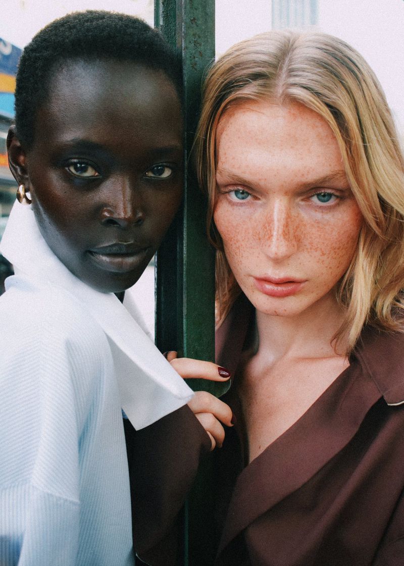 Two models pose closely together, one with dark skin and a light blue shirt, the other with fair skin and a brown jacket, in an outdoor urban setting.